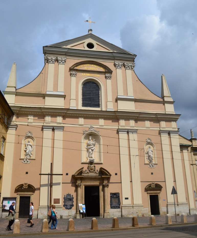 Współczesne zdjęcie przedstawiające barokową fasadę kościoła ojców karmelitów na Piasku w Krakowie. Fasada jest dwukondygnacyjna, rozgraniczona architrawem i gzymsem. Dolne lico fasady dzielą pasy gładkich pilastrów z toskańskimi kapitelami. Górna część fasady posiada przy dolnej krawędzi gładki pas – cokół. Ta wyższa kondygnacja zamknięta jest po bokach obeliskami. Szczyt wieńczy przyczółek z okrągłym okienkiem. Na osi budynku pojawiło się okno z arkadowo zamkniętym ościeżem, nad nim półokrągły przyczółek, dźwigany przez dwa delikatne pilasterki. W nim napis przypominający o renowacji zakończonej w 1939 r. Do kościoła prowadzą trzy wejścia przy głównej elewacji.