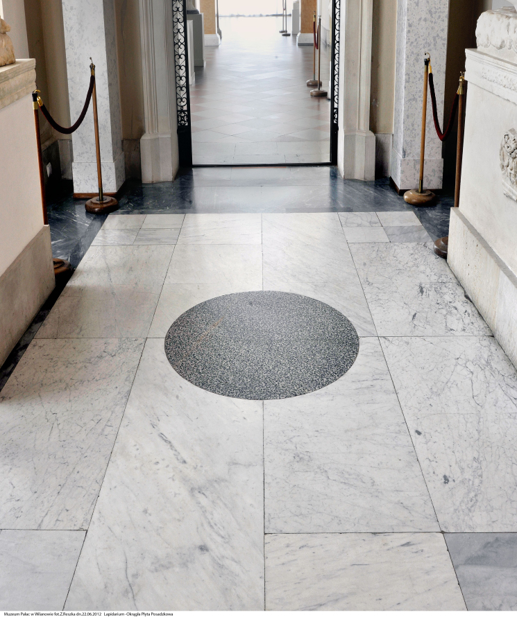 Zdjęcie przedstawia korytarz w pałacu. Na pierwszym planie posadzka z okrągłym, szarym elementem kamiennym. Jest to najcenniejszy fragment posadzki w Lapidarium.