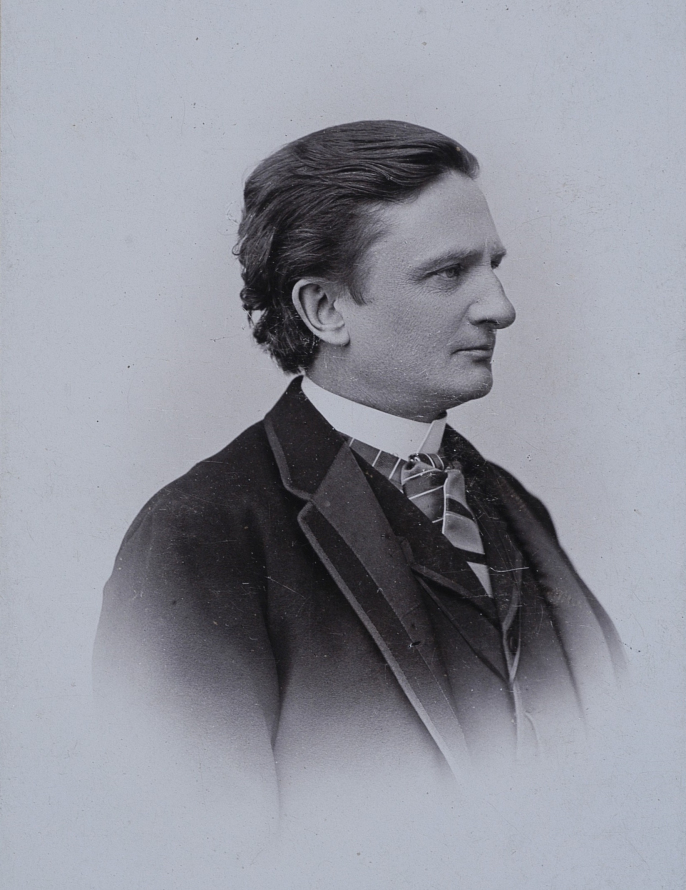 Portret mężczyzny z profilu. Mężczyzna ubrany jest w garnitur, kamizelkę białą, koszulę i krawat. Ma krótkie zaczesane do tyłu włosy. Jest zwrócony w prawo. Jest to portret Wincentego Rapackiego. 